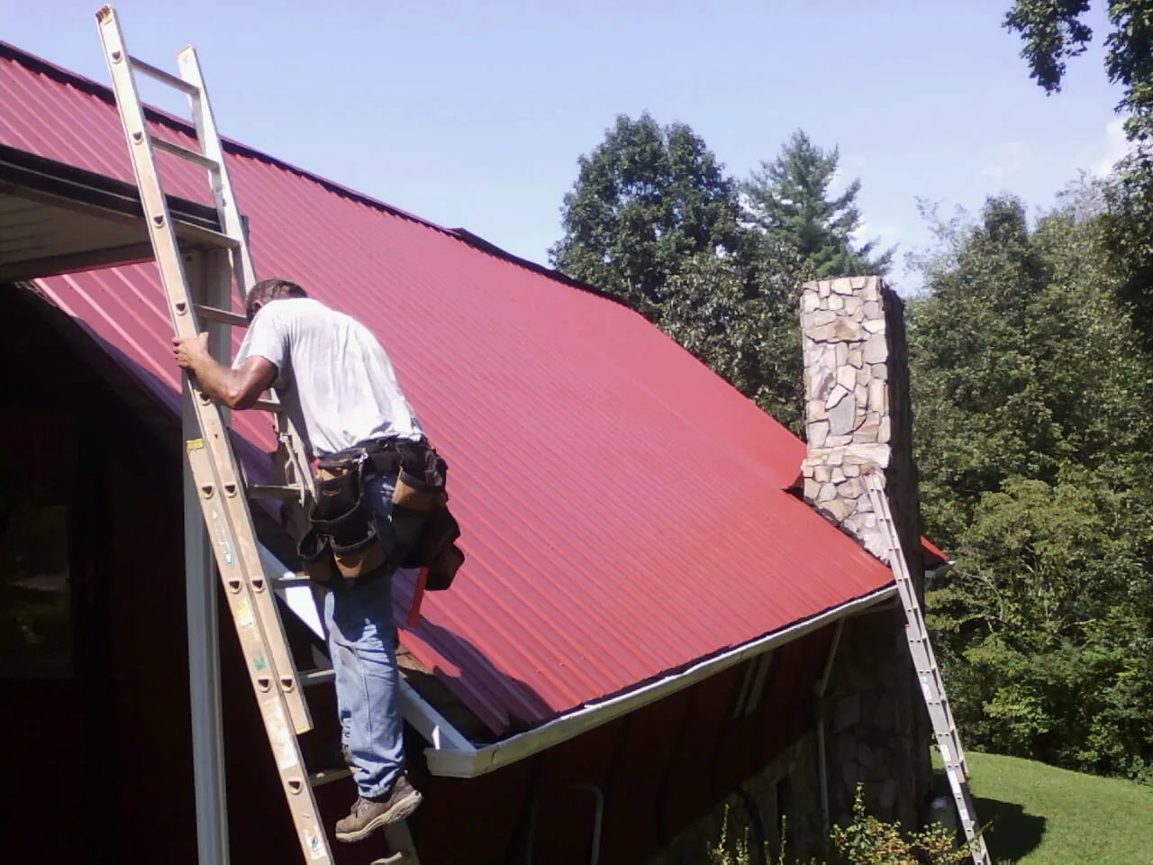 Roofer on a ladder beside a red metal roof and stone chimney — Soffit & Fascia Repair by Roofing Brothers in North Creek